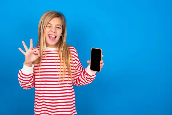 エキサイティングな美しい慎重な十代の少女は スマートフォンの空白の画面を示す青いスタジオの背景にストライプのシャツを着て 目を点滅し 手でOkのサインを行います 広告コンセプト — ストック写真