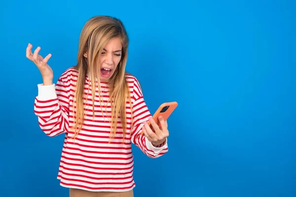 Photo of outraged annoyed beautiful caucasian teen girl wearing striped shirt over blue studio background holds cell phone, makes call, argues with colleague,  expresses negative emotions. People and anger.