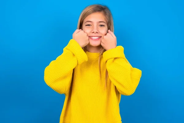 青い壁の上に黄色いセーターを着ている幸せな美しいブロンドの十代の少女は頬を広く笑わせ 良い気分にある肯定的な表現を持っています — ストック写真