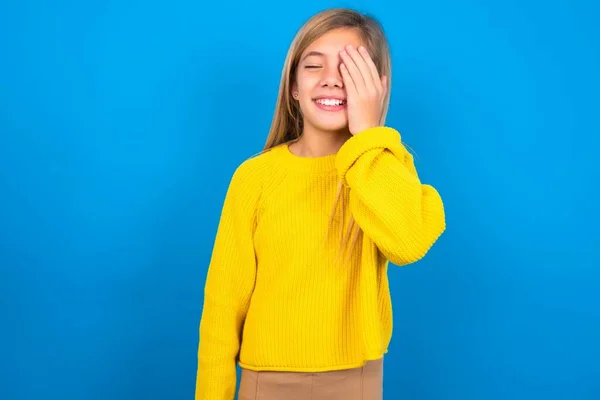 青い壁の上に黄色いセーターを着ている美しいブロンドの十代の少女は顔のヤシと笑顔を広くします 笑いは積極的に面白いジョークのポーズを聞きます — ストック写真