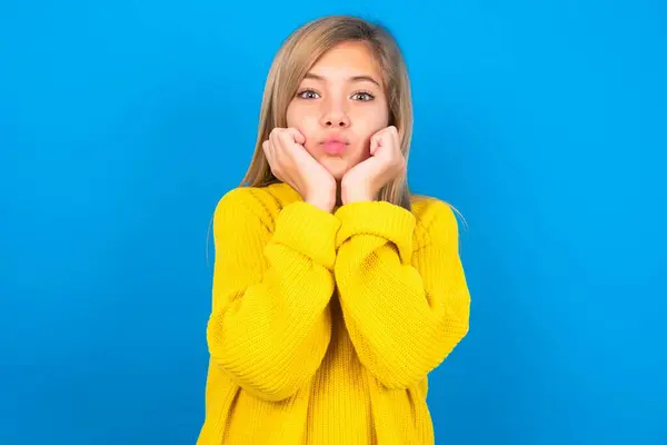 美しいブロンドの十代の少女は青い壁の上に黄色のセーターを着用し 驚いた表情は 唇を折り畳み続け 面白いグリムを作ります — ストック写真