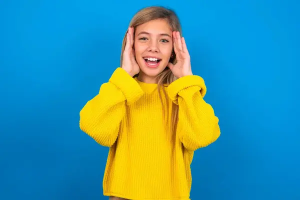 美しいブロンドの十代の少女は青い壁に黄色のセーターを着ています 陽気で 幸せな反応 — ストック写真