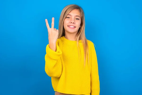 美しいブロンドの十代の少女は青い壁に黄色いセーターを着て笑顔でフレンドリーに見て 手を前方に2番目に示し カウントダウン — ストック写真