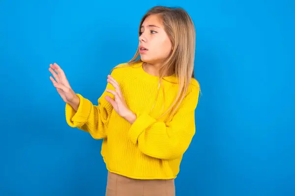青い壁の上に黄色いセーターを着ている不快な美しいブロンドの十代の少女は 空のスペースに向かって手を保ち より近づかないように求めます不快な何かを見ます — ストック写真