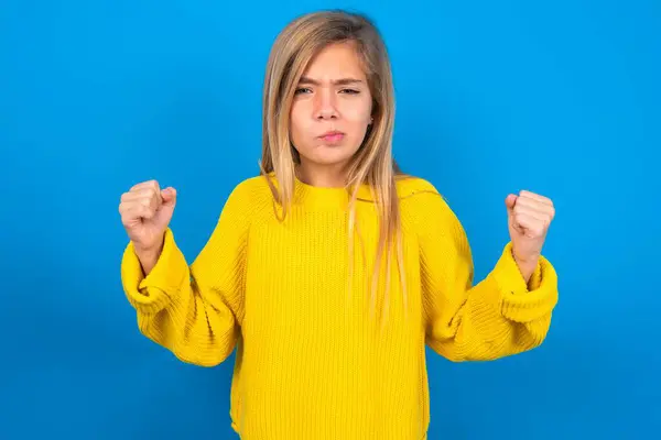 青い壁の上に黄色いセーターを着用した美しいブロンドのティーンガールは 怒りで頬を上げ レンチされた拳を上げる怒りと攻撃的な感情を表現します フューリアス モデル — ストック写真
