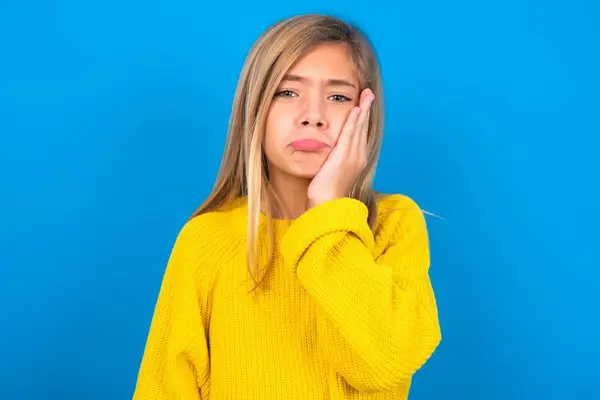 悲しい孤独な美しいブロンドの十代の少女は青い壁の上に黄色いセーターを着用し 手は唇を噛み 不快に目を向けます 悪い感情について — ストック写真