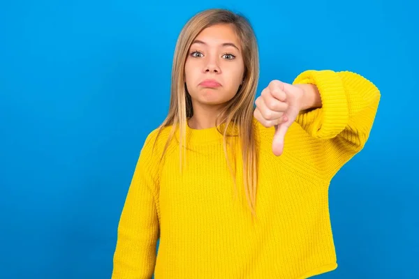 青い壁の上に黄色いセーターを着ている美しいブロンドの十代の少女は 不幸で怒って 指の下のジェスチャーで拒絶と否定を示しています バッド表現 — ストック写真