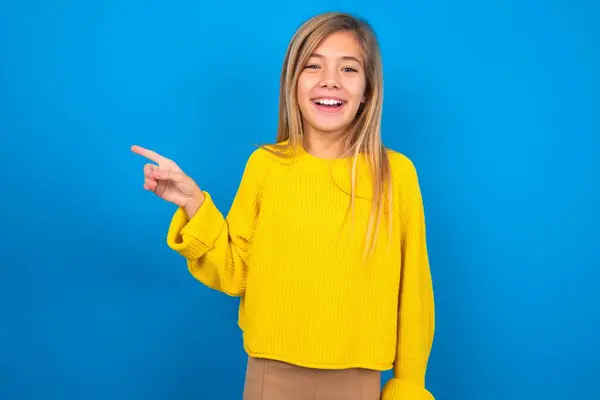 青い壁の上に黄色いセーターを着ている美しいブロンドの十代の少女は幸せに空白のスペースを指摘し 良いニュースや予期しない販売に興奮ショッピング割引オファーを示しています — ストック写真