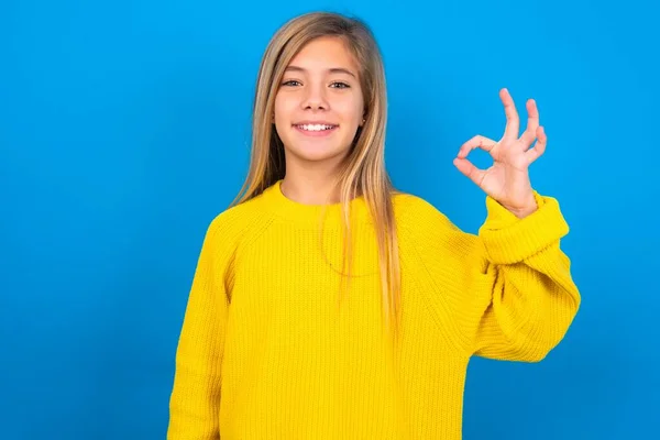 青い壁の上に黄色いセーターを身に着けている美しいブロンドの十代の少女は手の腕のオキーのシンボルを承認し 新鮮なニュースを助言します — ストック写真