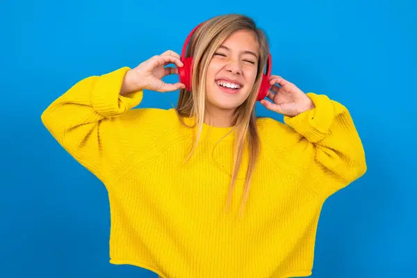 beautiful blonde teen girl wearing yellow sweater over blue wall smiles broadly feels very glad listens favourite music track via wireless headphones closes eyes.