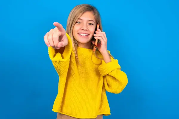 青い壁に黄色いセーターを着用したポジティブな美しいブロンドの十代の少女は カメラで直接電話会話の笑顔を広く楽しんでいることを示しています あなたは私に参加する — ストック写真