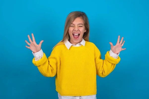 青い壁の上に黄色いセーターを着ている感情的な慎重な十代の少女は大声で笑い 面白いジョークや物語を聞き 満足して手のひらを上げます — ストック写真