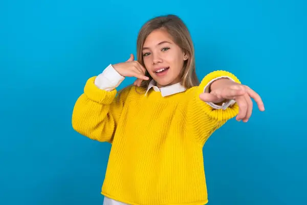 caucasian teen girl wearing yellow sweater over blue wall smiling cheerfully and pointing to camera while making a call you later gesture, talking on phone