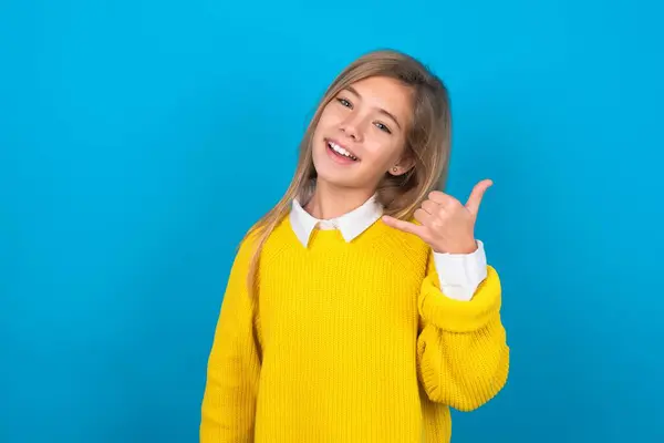 caucasian teen girl wearing yellow sweater over blue wall smiling doing phone gesture with hand and fingers like talking on the telephone. Communicating concepts.