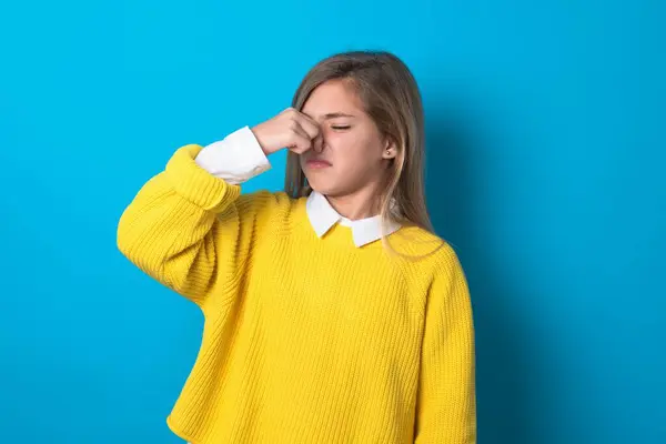 青い壁の上に黄色いセーターを着ている白人の十代の少女は臭いと嫌な 耐え難い臭いを嗅ぎ 鼻に指で息を止めます 悪い匂い — ストック写真