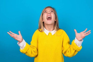 Mavi duvarın üzerinde sarı kazak giyen beyaz bir kız. Deli gibi bağırıyor, agresif bir ifadeyle bağırıyor ve kollarını kaldırıyor. Hayal kırıklığı konsepti.