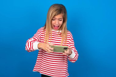 Mavi duvarın üzerinde çizgili tişört giyen heyecanlı sarışın bir kızın portresi cep telefonuyla oyun oynuyor..