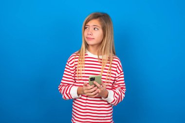 Mavi duvarın üzerinde çizgili tişört giyen sarışın genç kız. Telefondan el ele tutuşmuş. İyi gençlik haberleri okuyor. Boş yer ilanı gibi.