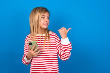 Sarışın genç kız mavi duvarın üzerinde çizgili tişört giyiyor. Akıllı telefonuyla mesaj atıyor ve gülümseyen bir gülümsemeyle baş parmağıyla işaret ediyor.