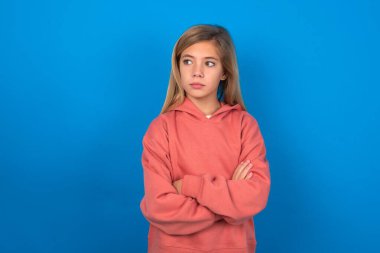 Mavi duvarın üzerinde pembe kazak giyen güzel, düşünceli, beyaz bir kız kollarını kavuşturmuş, düşünceli bir ifadeyle ne yapacağını düşünüyor.