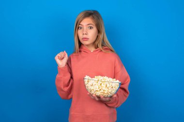 Şaşırmış duygusal açıdan güzel beyaz kız mavi duvarın üzerine pembe kazak giyip avuçlarını ovuyor ve inançsızlıkla kameraya bakıyor.