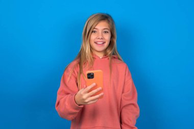 Mavi duvarda pembe kazak giyen güzel beyaz kız cep telefonunu elinde tutuyor ve olumlu haberler veriyor, modern cep telefonu kullanıyor.