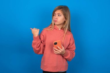 Mavi duvarın üzerinde pembe kazak giyen güzel beyaz kız başparmağıyla işaret ediyor ve boş alan gösteriyor, cep telefonunu mesaj göndermek için tutuyor..