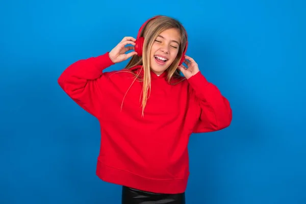 blonde teen girl wearing red sweater over blue wall smiles broadly feels very glad listens favourite music track via wireless headphones closes eyes.
