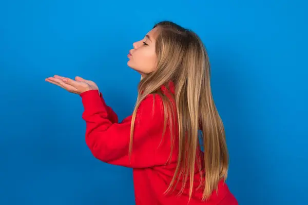 プロフィールの側面の眺め 魅力的なブロンドの十代の少女の肖像画は青い壁に赤いセーターを着て空気のキスを送ります — ストック写真