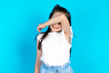 Mavi stüdyo arka planında duran güzel beyaz kız. Gözleri kollarıyla kaplıyor. Neşeli ve komik gülümsüyor. Kör kavram.