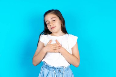 Mavi stüdyonun arkasında duran güzel beyaz kız elleri göğsünde, gözleri kapalı ve yüzünde minnet dolu bir jest ile gülümsüyor. Sağlık kavramı.