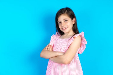 Mavi arka planda pembe elbiseler giyen, kollarını kavuşturup kameraya gülümseyen güzel bir kız. Pozitif kişi.