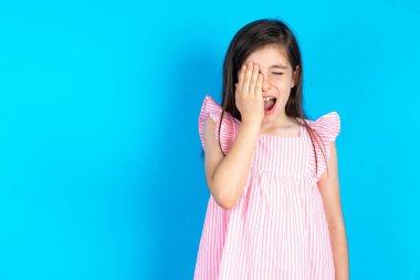 Mavi arka planda pembe elbise giyen güzel bir kız çocuğu esnemekten yoruldu. Yüzünün yarısını, gözünü ve ağzını eliyle kapatmaktan yoruldu. Yüzüm acıyor..