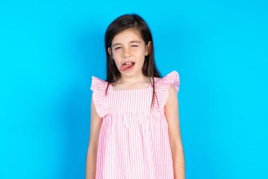 Mavi arka planda pembe elbiseler giyen güzel bir kız dilini komik bir ifadeyle dışarı çıkartıyor. Duygu kavramı.