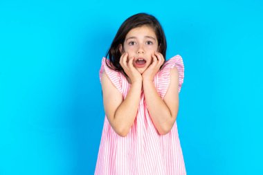 Sersemlemiş, mavi arka planda pembe elbise giyen güzel bir kız çocuğu heyecan ve heyecanı ifade eder, çenesini indirir, elleri yanaklarında, gözleri fırlar