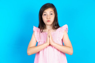 Pozitif çocuk mutlu bir şekilde gülümsüyor. Müdahaleden hoş haberler aldığına seviniyor. Avuç içlerini bir arada tutuyor. Güzel beyaz kız mavi stüdyo arka planında poz veriyor.