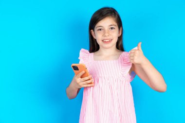 Akıllı telefonuyla mesajlaşan gülümseyen çocuğun portresi tamam işareti yapıyor, başparmağını parmaklarıyla kaldırıyor, mükemmel bir işaret. Güzel beyaz kız mavi stüdyo arka planında poz veriyor.