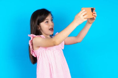 Sosyal medyaya göndermek için selfie çeken bir çocuk ya da arkadaşlarıyla video görüşmesi yapan bir çocuk. Güzel beyaz kız mavi stüdyo arka planında poz veriyor.