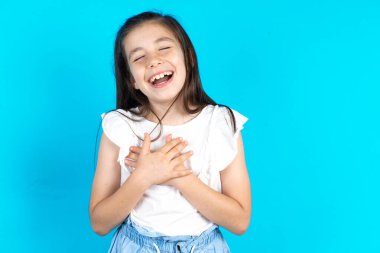 Çocuk mutluluğu ifade ediyor, güzel gülüyor, ellerini kalbinden ayırmıyor. Güzel beyaz kız mavi stüdyo arka planında poz veriyor.