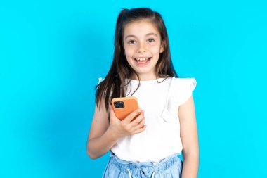 Çocuk cep telefonunu elinde tutuyor ve olumlu haberler veriyor, modern cep telefonu kullanıyor. Güzel beyaz kız mavi stüdyo arka planında poz veriyor.