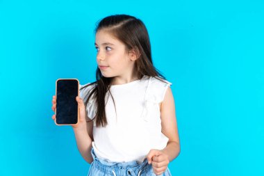 Çocuğun elinde yeni bir cep telefonu var ve gizemli görünüyor. Modern cep telefonunun boş görüntülerini gösteriyor. Güzel beyaz kız mavi stüdyo arka planında poz veriyor.