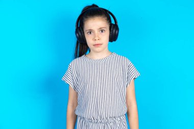 Ciddi şekilde canı sıkkın bir çocuk kameraya kızmış, müzik kulaklığı takıyor, müzik dinliyor. Çizgili tişörtlü güzel beyaz kız mavi stüdyo arka planında duruyor. 