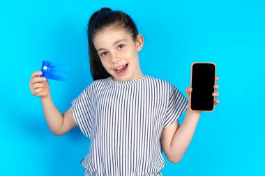 Çocuk banka hesabı açmış, elinde akıllı telefon ve kredi kartı var, gülümsüyor, online alışveriş başvurusu öneriyor. Çizgili tişörtlü güzel beyaz kız mavi stüdyo arka planında duruyor. 