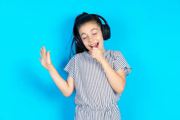 Happy kid sings favourite song keeps hand near mouth as if microphone wears wireless headphones, listens to music. Beautiful caucasian girl in striped dress standing over blue studio background