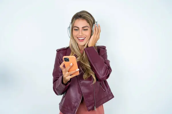 Joyous young caucasian woman wearing leather jacket   poses with mobile phone device, types text message on modern smartphone, watches funny video during free time, enjoys good internet connection,
