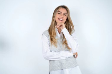 Optimistic caucasian woman wearing white shirt keeps hands partly crossed and hand under chin, looks at camera with pleasure. clipart