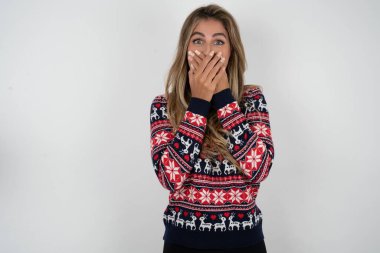 Şaşkın Kafkasyalı kadın Noel süveteri giyiyor. İki eli de ağzında, hayret verici bir şeyden korkuyor.