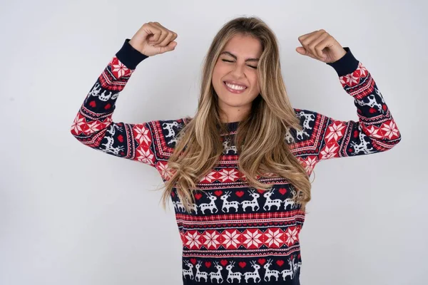 Strong powerful caucasian woman wearing christmas sweater toothy smile, raises arms and shows biceps. Look at my muscles!