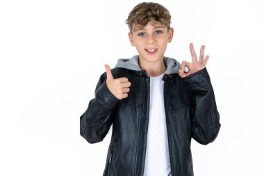 Beyaz stüdyo arka planındaki yakışıklı beyaz çocuk gülümsüyor ve mutlu görünüyor, gamsız ve pozitif, tek elle zafer ya da barış işareti yapıyor.