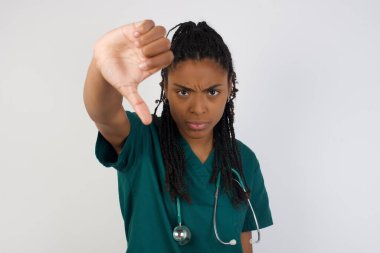 Hoşnutsuz Afrikalı Amerikalı doktor kadın onaylamadığını, başparmağını aşağıda tuttuğunu, hoşlanmadığını ifade ettiğini, hoşnutsuzca kaşlarını çattığını, gri arka planda soyutlandığını gösteriyor. Vücut dili kavramı.